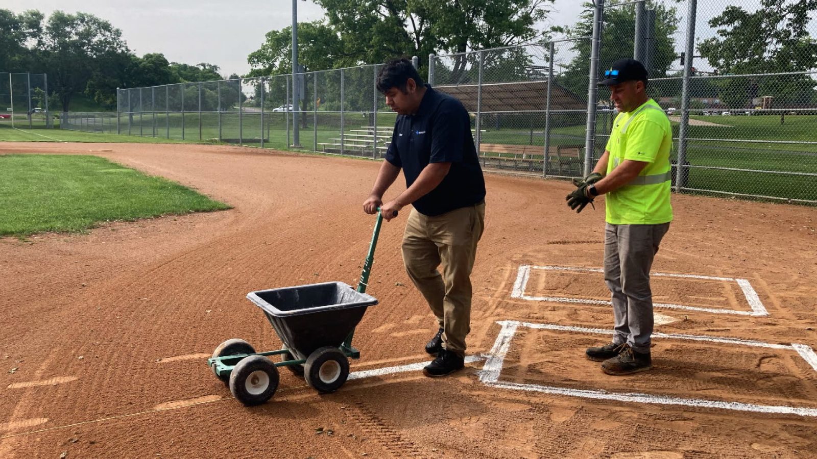 Young man with a chalking device spreads lines on a baseball diamond while a city employee supervises