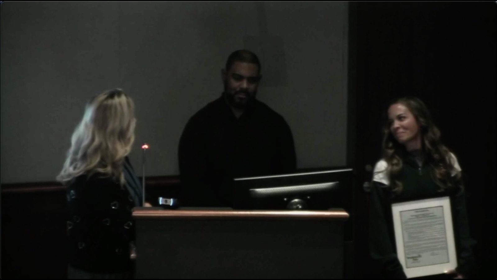 Two people stand at a podium beside Juaire, holding a plaque