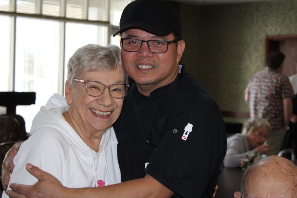 filipino man in a black outfit embracing an elderly white woman wearing a white hoodie